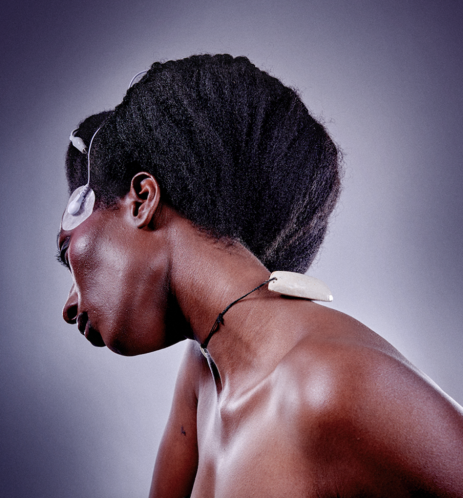 Chienne de Garde/Vanessa Sindihebura est une personne afrodescendante, elle pose devant un fond gris, son afro est coiffé dans un grand chignon, elle porte un collier avec un pendentif blanc. Collé à sa tempe, une électrode blanc. Elle regarde elle tourne la tête et a le regard rivé vers le sol.