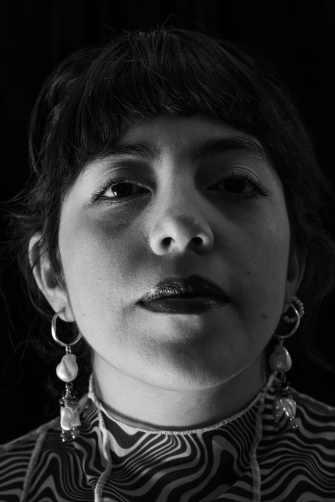 Cecilia regarde face à la caméra, la tête haute. Elle se tient devant un fond noir, et la photo est en noir et blanc. Cecilia poste des boucles d'oreille avec une perle pendante.