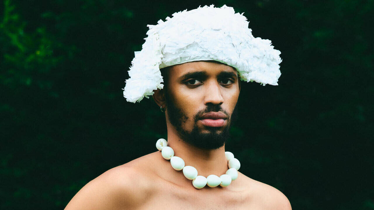 Marvin M'Toumo, afrodescendant pose devant la caméra. Il a un regard sérieux et porte un chapeau fait avec un matériau qui rappelle des plumes qui ressemble à un bicorne (chapeau napoléonien), ainsi qu'un collier à grosses perles blanches.