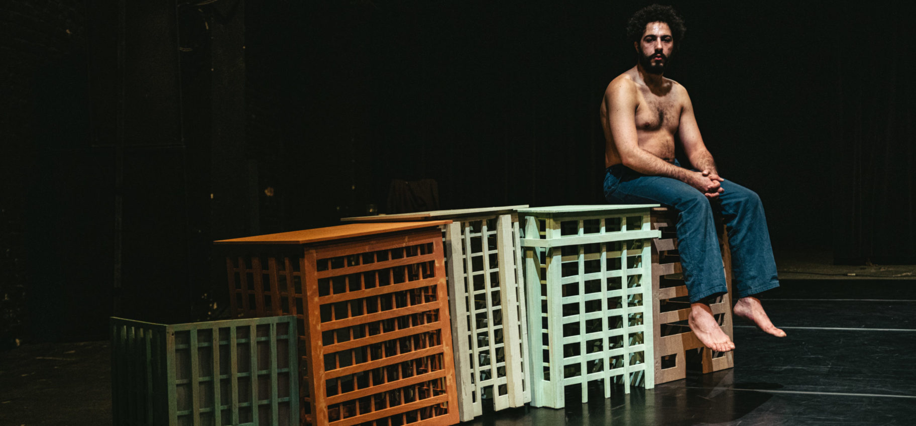 Photo du spectacle. Habib Ben Tanfous porte un pantalon bleu. Il est torse et pieds nus. Il est assis sur un décor composé d'immeubles miniatures. Ses épaules sont baissées, ses mains sont jointes et reposent sur ses cuisses. Son expression est pensive.