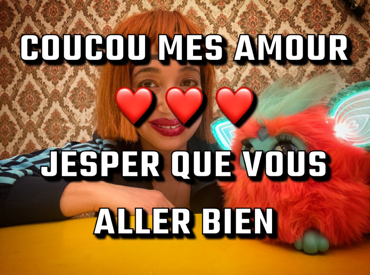 Une photo de Lamya Moussa avec un texte mal orthographié qui dit: Bonjour mes amours, coeur coeur coeur, j'espère que vous allez bien. Lamya sourit largement, et porte une perruque rousse, un jouet Furby poilu à ses côtés.