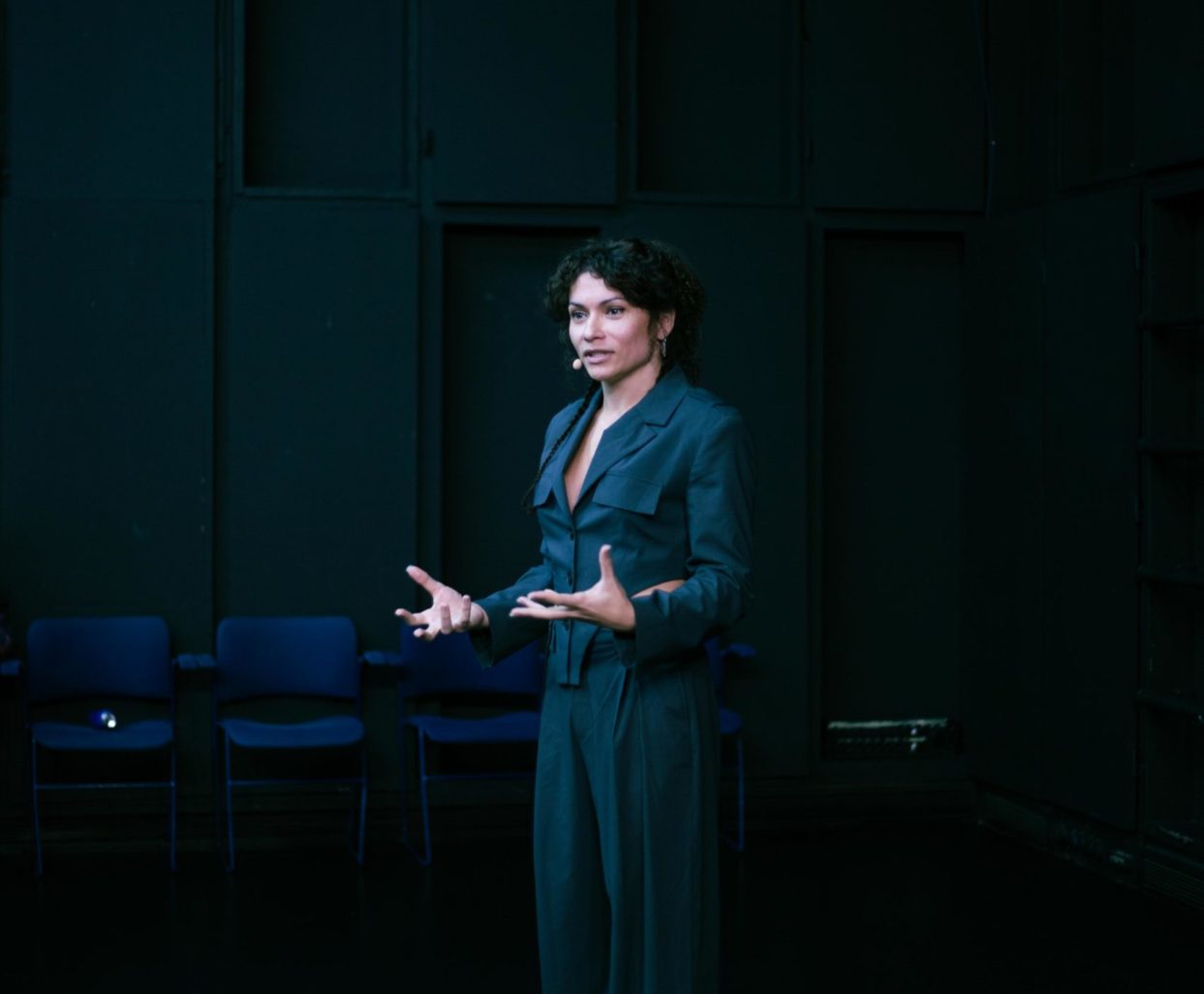 Gaya est debout sur une scène apparemment vide et s'adresse au public. Elle a les cheveux bruns, longs et bouclé, accrochés en chignon derrière sa tête. Elle porte un ensemble gris foncé, très classe.
