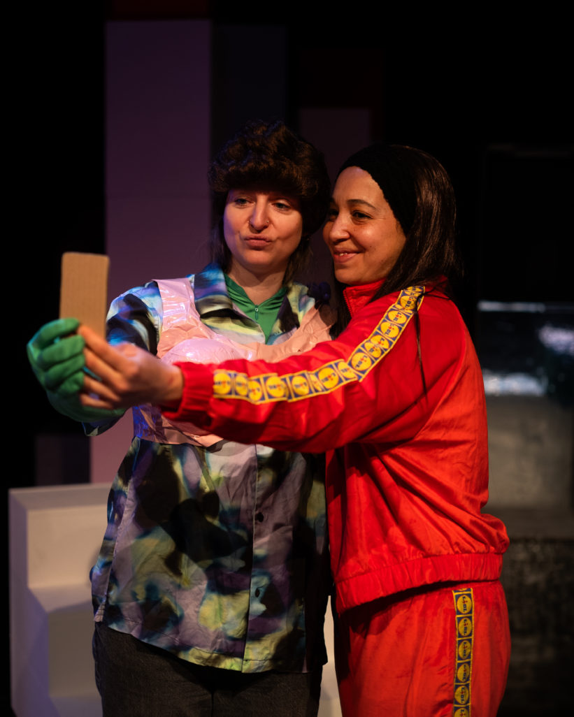 Photo de Claire Forclaz et Lamya Moussa qui se prennent ensemble en selfie. Claire porte des énormes faux seins en plastique gonflable par dessus une chemise aux manches courtes. Ses bras sont recouverts de gants en élasthane vert. Elle porte une perruque type coupe mulet. Elle fait une duckface. Lamya porte un survêtement de sport rouge. Les bandes latérales du survêtement sont le logo du supermarché Liddl. Elle sourit en regardant le téléphone. Les deux artistes tiennent à bout de bras ce téléphone qui est en fait un carton découpé à la taille d’un smartphone.