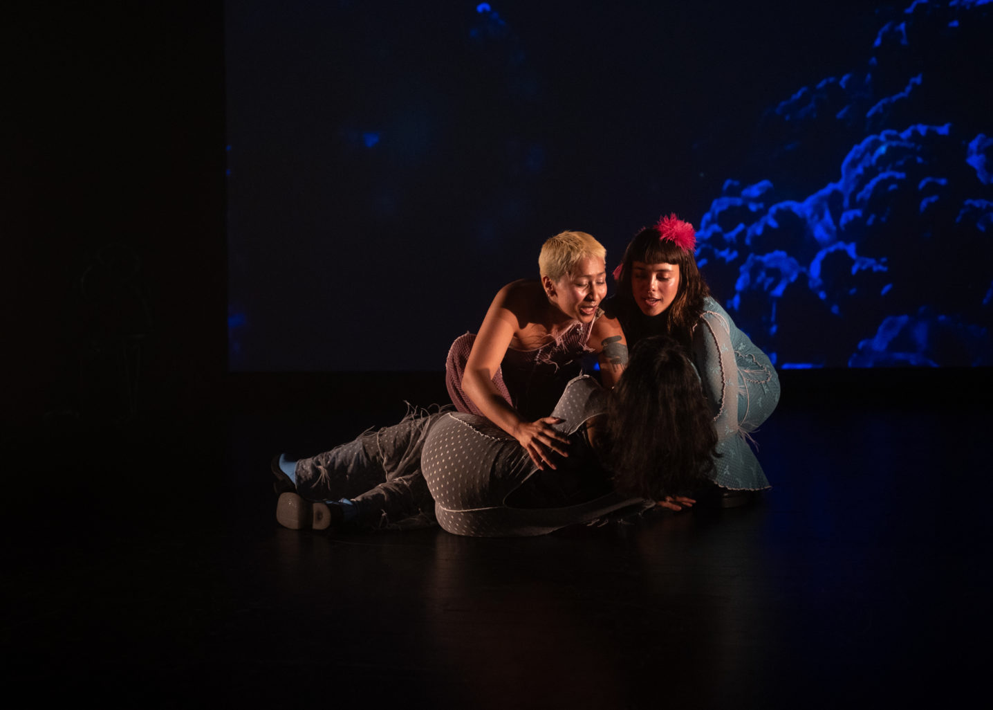 Au fond de scène, un écran projette une image abstraite de nuages bleus. Au centre de la scène, dans une lumière chaude, deux personnages soutiennent un personnage couché de dos. Elles semblent vouloir la rassurer.