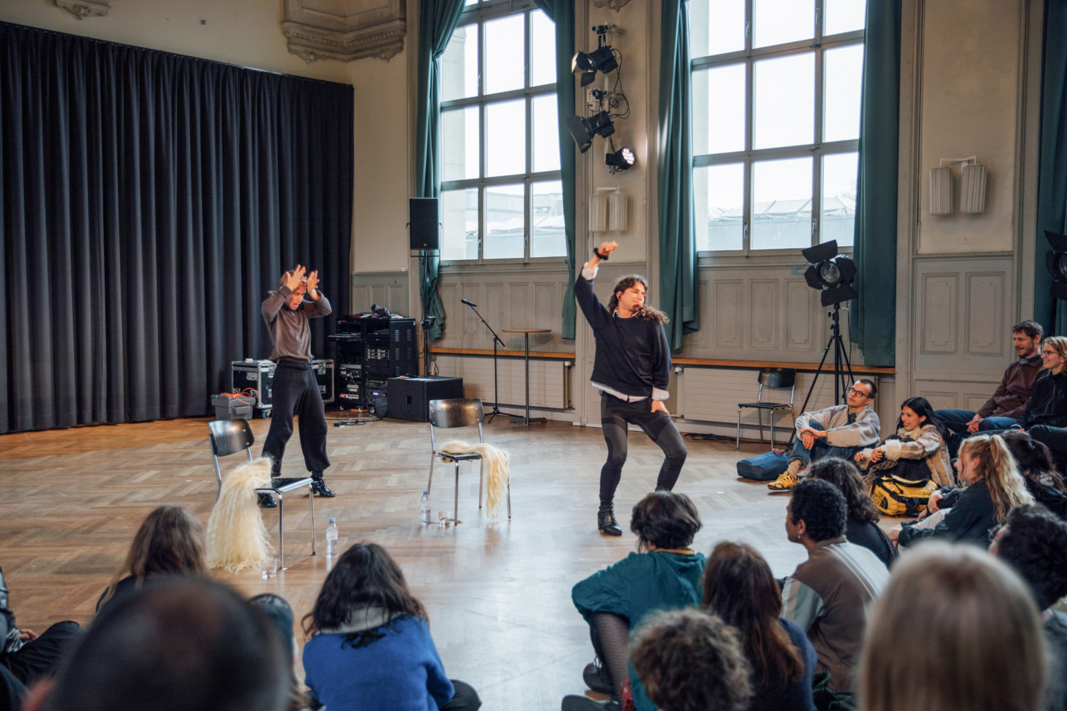 Dans une salle de spectacle sans gradin, deux personnes, l'une blonde au cheveux court, l'autre brune au cheveux longs et lisses sont debout en train de performer en faisant des poses dramatiques. L'une lève ses deux grande ouverte posée sur sont front et l'autre semble se balancer en pliant les jambe et levant son bras droit. A coté d'ielles, deux chaises noirs sur lesquelles sont posées deux perruques blondes aux cheveux longs. face à elle un public assis en demi-cercle au sol sur un parquet en bois.