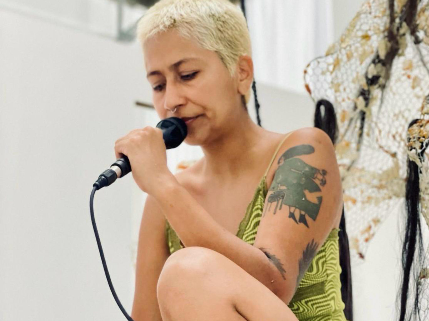 Dans une salle claire, Valeska est assise au premier plan et occupe presque toute l'image. Elle a les cheveux courts blonds décolorés, a des tatouages visibles et porte un short en jeans et un top vert clair qui ne couvre pas ses épaules. Assise avec un genou au sol et l'autre replié en auteur, elle tient dans sa main un micro avec fil et ferme les yeux tout en parlant dans celui-ci.