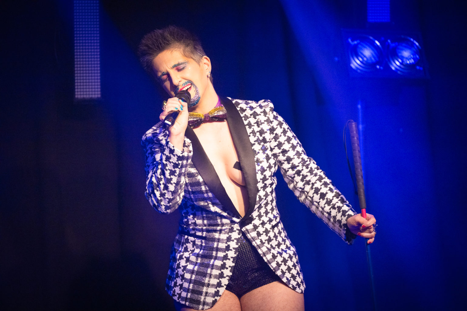 Tito Bone, drag king est sur scène, il chante en tenant un micro. Dans l'autre main, il tient sa canne blanche. Il a les cheveux courts, les sourcils et une moustache pailletés. Il a un noeud papillon noir autour du cou et un blazer à carreaux noir et blanc ainsi que des shorts très courts et noirs. Derrière lui il y a de la lumière bleue.