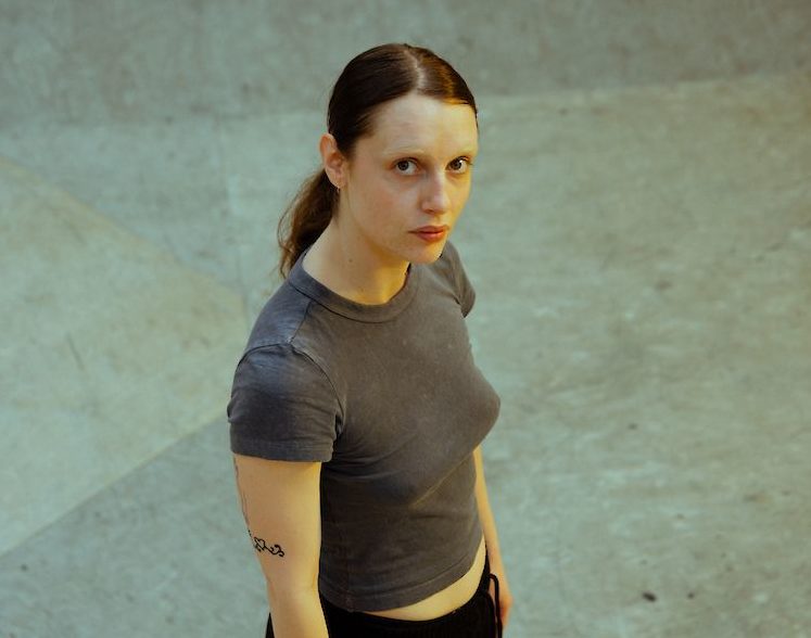 lisa laurent, chorégraphe et interprète porte une t-shirt gris, un bas de training noir. lisa a les cheveux brun attaché, un tatouage sur son bras droit et lisa lève les yeux vers l'objectif de l'appareil photo.