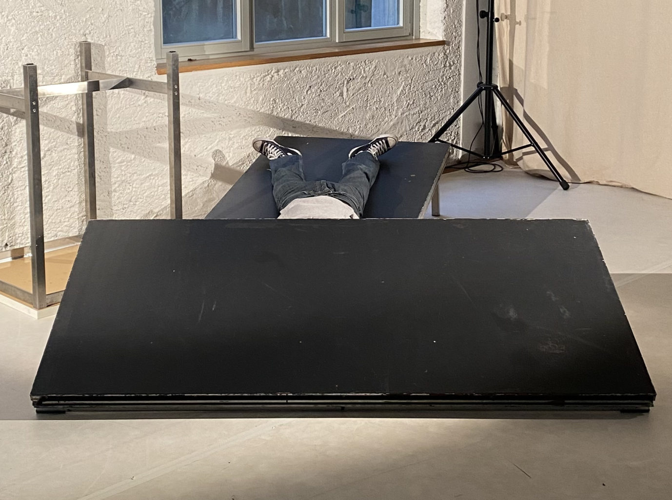 Dans un espace bien éclairé, une personne est allongée sous un décor qui s'écroule. On ne perçoit qu'un bout d'un top blanc et un jeans bleu foncé. La personne est couchée sur une blanche noire à la verticale alors qu'une autre planche noire horizontale l'ensevelie. Il y a une grande vitre en arrière plan et un haut parleur sur pilotis.