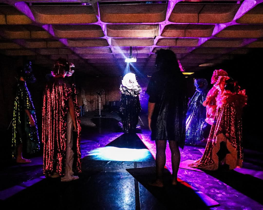 Septs personnages habillées de masques et drapés brillants et flamboyants sont en cercle debout. Un des personnages est illuminé d'une lumière blanche, il est face à nous. Il fait sombre dans la salle.