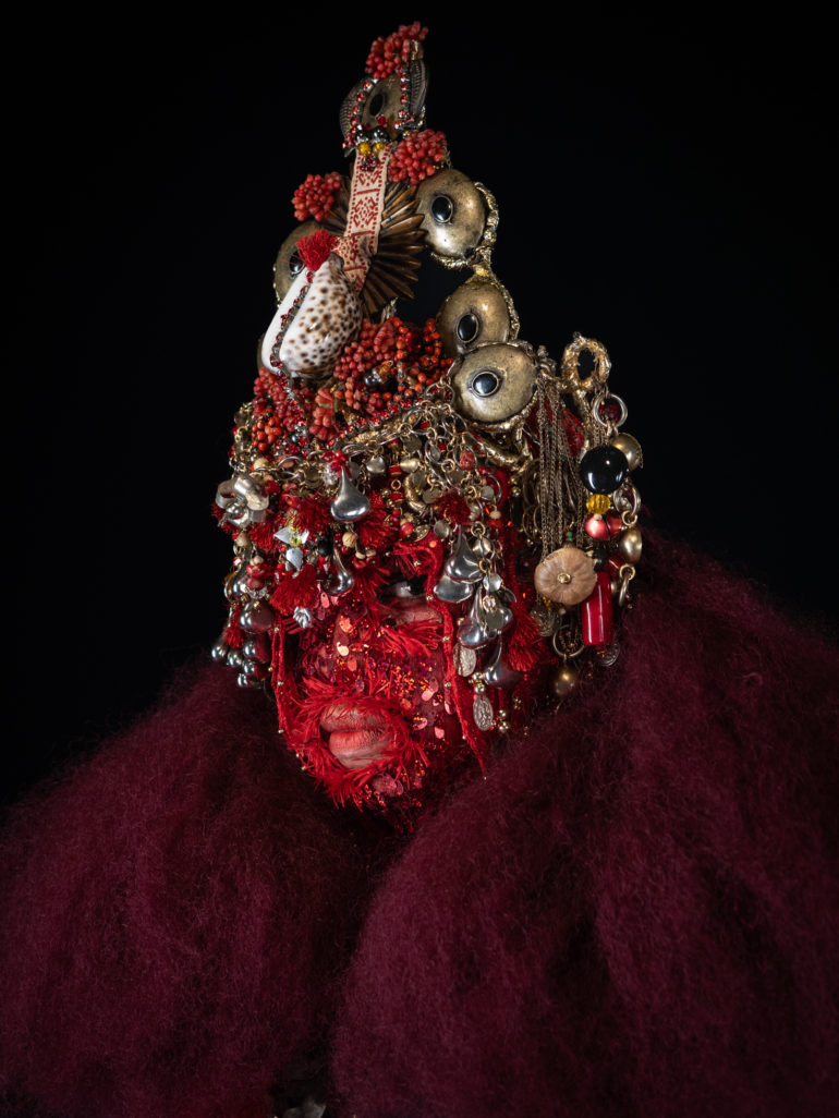 Transälien pose avec un masque rouge, orné de bibelots en or et argent, ainsi que de coquillage. Le masque est pointu et majestueux. Elle porte aussi un manteau bordeaux en feutre en regardant droit dans l'objectif de la caméra, dans un fond noir.
