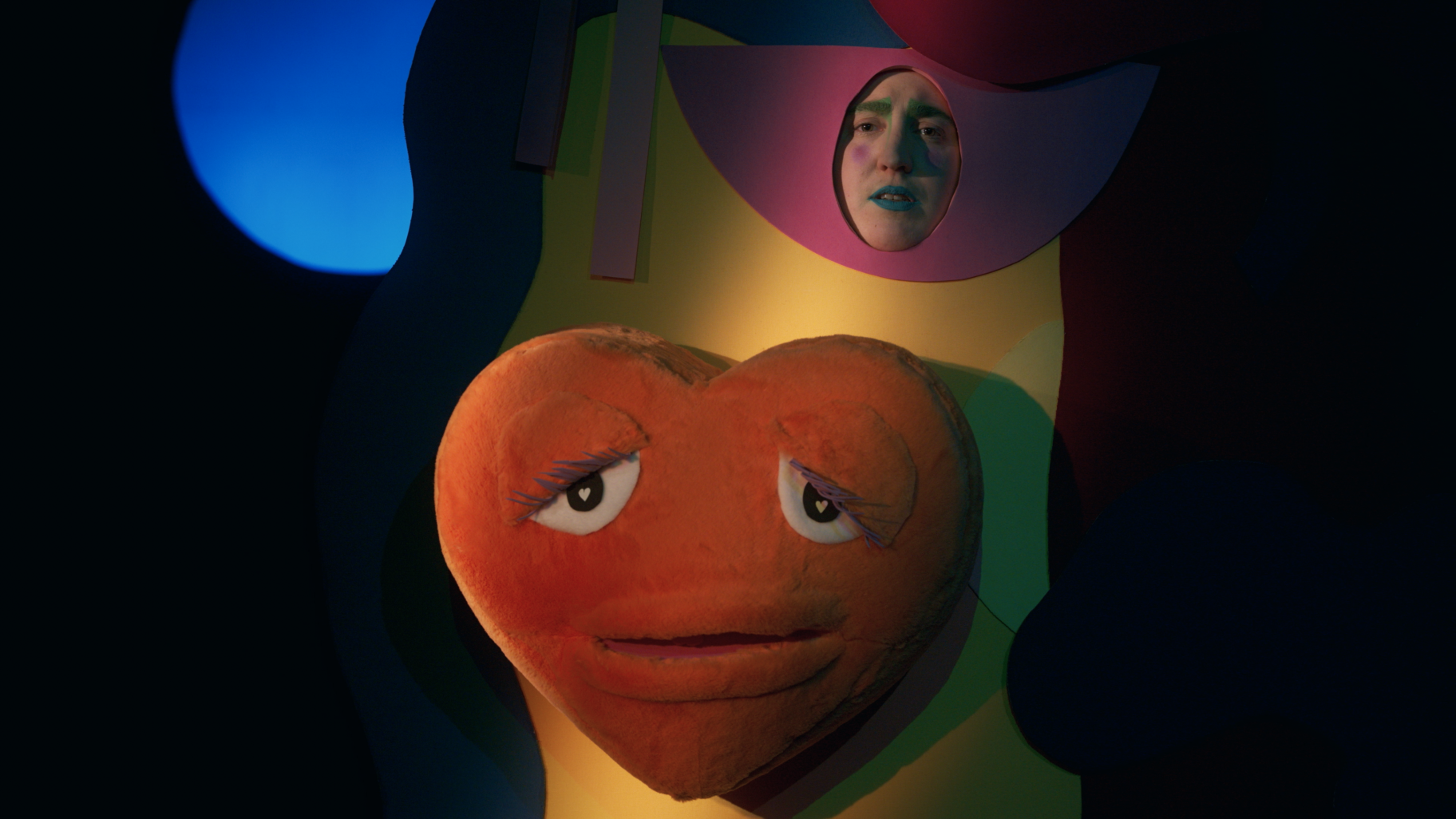L'image montre l'artiste Louise Siffert dans un costume jaune avec une énorme peluche en forme de coeur au centre du costume. Attaché au costume est un masque rose en forme d'oeil qui ne laisse apercevoir que le visage de Louise. Son visage est maquillé avec les sourcils verts, les joues rose et les lèvres bleues. Louise a l'air triste.