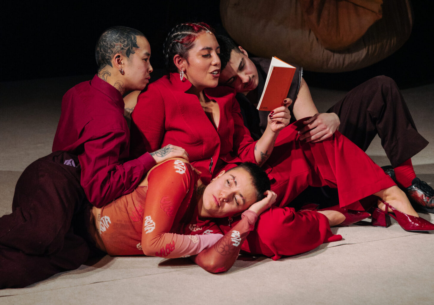Cecilia Moya Rivera est posée au sol, entrelacée par 3 de ses protagonistes. Tout le monde est vêtu de rouge. Cecilia lit un poème provenant de son livre rouge. La scène est bien éclairée et en arrière plan on aperçoit deux des sacs qui pendent depuis le plafond.