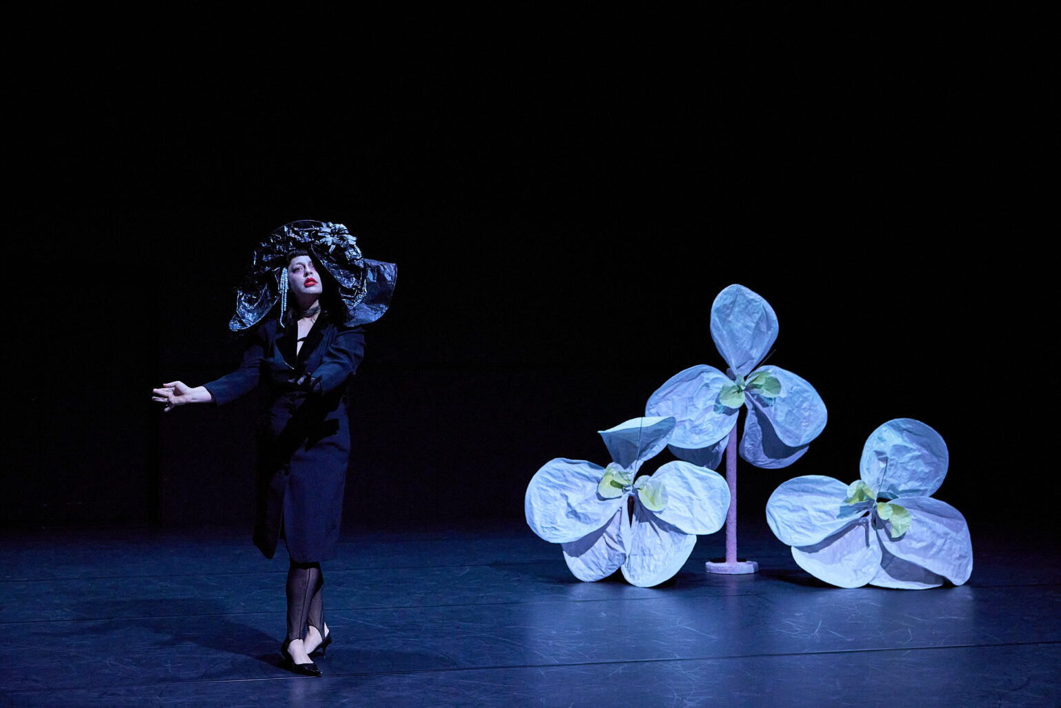 Marlène Charpentié se tient debout, la tête légèrement surélevée, vêtu de noir, maquillage simple accentué par un rouge pétant sur les lèvres. Elle gesticule ses mains, à côté d'elle des grosses fleurs en papier mâché. Marlène et les fleurs sont devant un fond noir.