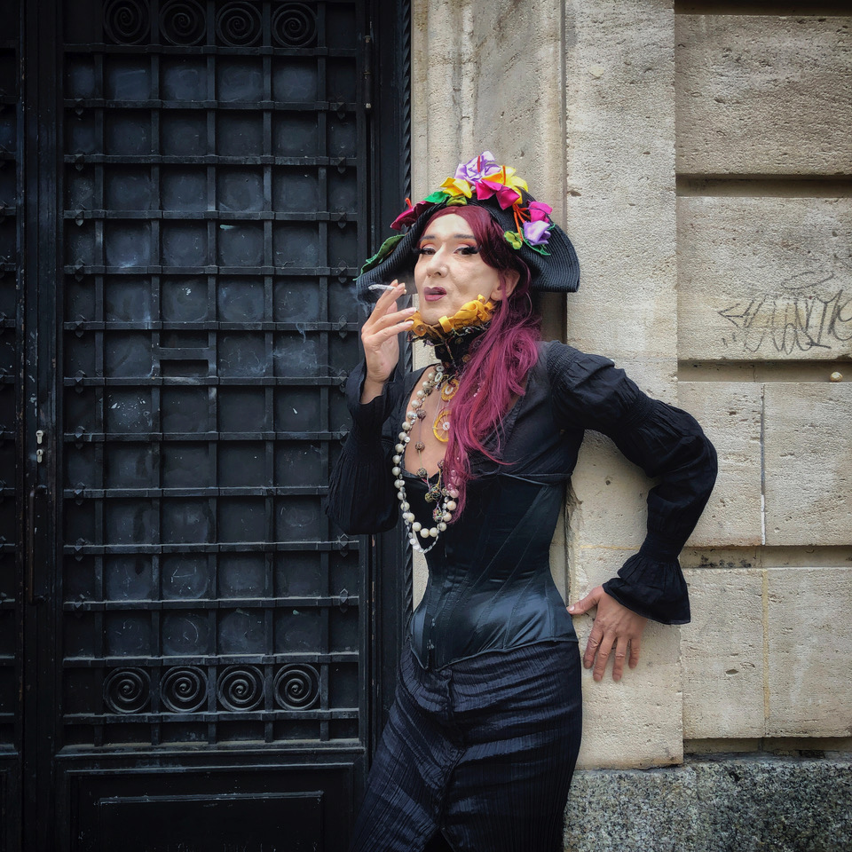 Greta est habillée d'une longue robe noire à corset, elle porte une bicorne noire décorée de fleures colorées. Elle a les cheveux violets attachés en une queue de cheval basse. Elle est dos contre le mur, devant la porte principale du centre autogéré de l'Usine, une main appuyée contre le mure alors que l'autre tient une cigarette consumée qu'elle approche près de sa bouche. Elle regarde du coin la caméra.