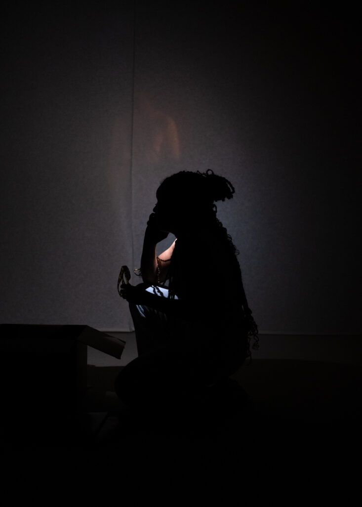 Elle, est accroupie dans le noir, le téléphone à la main. On en perçoit que sont ombre. En second plan, une silhouette légèrement illuminée se perçoit.