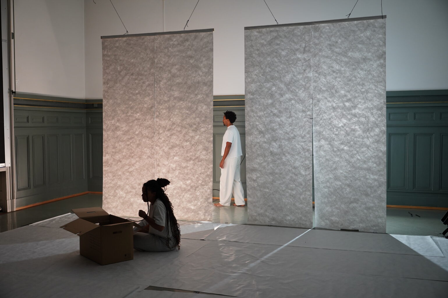 ELLE est assise dans l'ombre, LUI est en arrière-plan entre deux rideaux blanc. Les deux regardent le même mur.