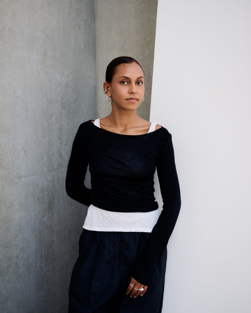 Portrait d'Aurélie Viviarié vêtue d'un pull à longues manches ajusté noir et un un sous pull blanc qui dépasse.à peine et d'une robe noir. Elle s'appuie sur un fond bétonné clair.